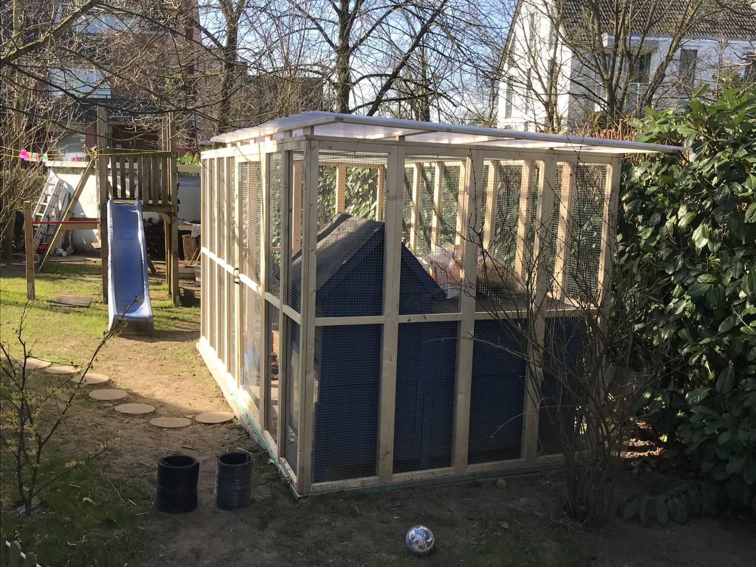 Neben einer Kelttergerüst für Kinder steht die Viliere für Kleintiere in einem Garten.