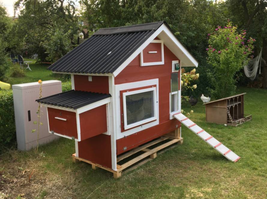 Ein angemaltes Statteldachhühnerhaus mit Außenlegenest und Rampe steht in einem gepflegten Garten.