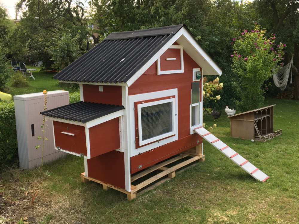 Ein angemaltes Statteldachhühnerhaus mit Außenlegenest und Rampe steht in einem gepflegten Garten.